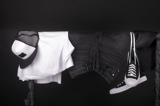 Hanging clothing. Black and white sneakers, cap and jeans on clothes rack on black.