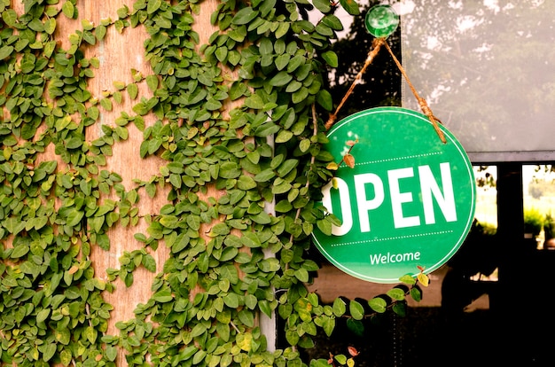 Hanging green sign with message OPEN on window with leaves ivy on wooden wall at coffee shop