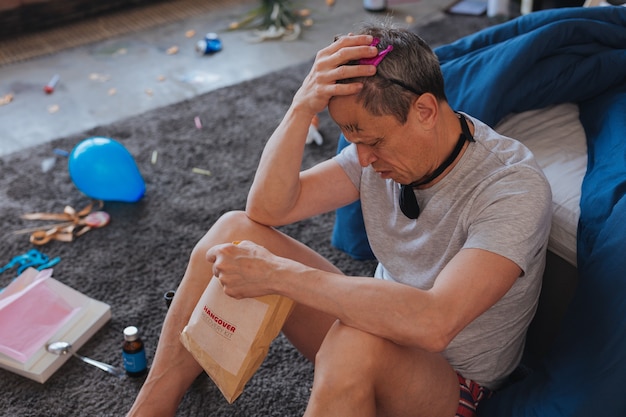 Hangover symptoms. Top view of fatigued mature man sitting on floor and studying recovery kit