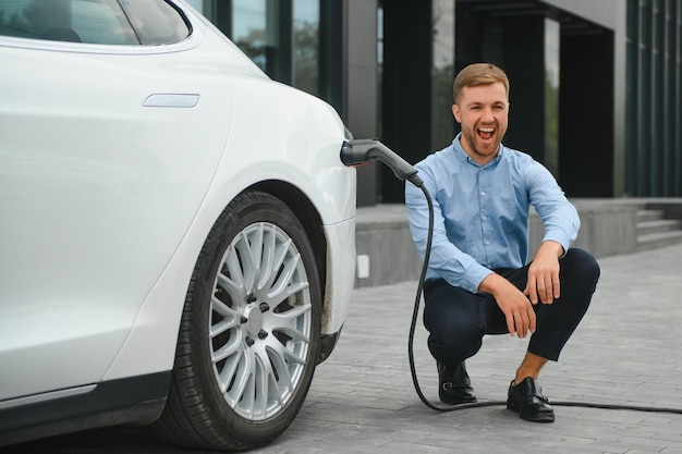 彼の新しい現代の電気自動車の近くに座って、車が充電ステーションで充電している間、充電器のプラグを持っているハンサムなひげを生やした男