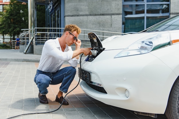 Hansome guy sitting near his new modern electric car and holding plug of the charger, while car is charging at the charging station