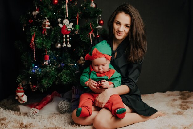 Happ mother sitting with baby little boy son at Christmas tree