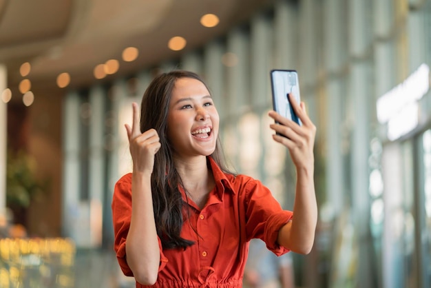 Счастье азиатская женщина улыбка рука машет рукой поцеловать семью через видеозвонок смартфон устройство социальное дистанцирование приветствие с размытым фоном торгового центра новый нормальный образ жизни