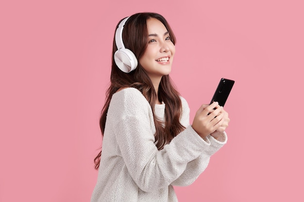 Happiness beautiful asian smiling young woman wearing\
technology wireless headphones for listening the music via smart\
mobile phone on pink background