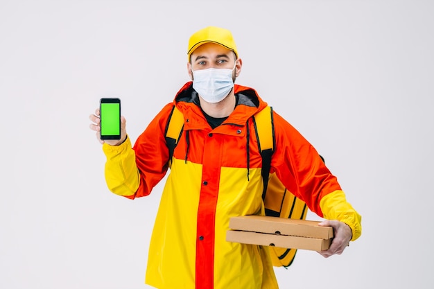 Fattorino della felicità che tiene la pizza che mostra il suo schermo verde dello smartphone che dà l'ordine indossando la maschera durante la pandemia di covid19 in studio su superficie bianca