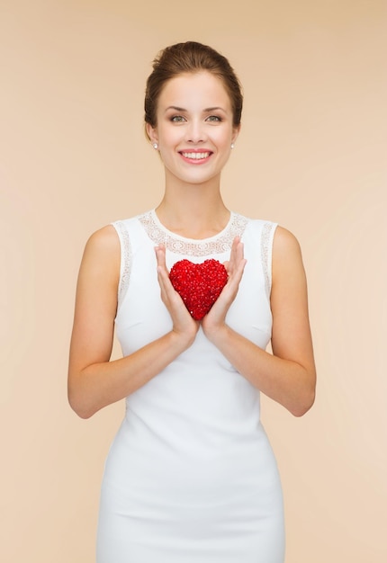 happiness, health, charity and love concept - smiling woman in white dress with red heart over beige background