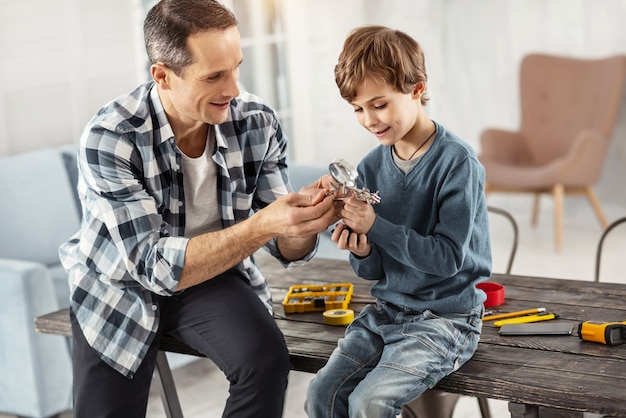 私の中の幸せ。テーブルに座っている間彼の息子と彼の近くに座っている彼の息子に楽器を見せている魅力的な幸せな黒髪の父