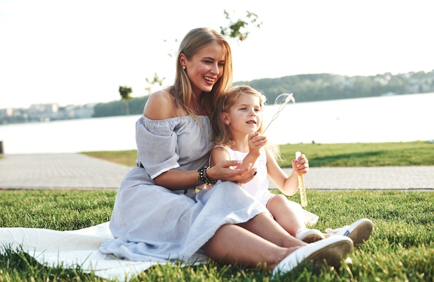 Happiness is in small things. Photo of young mother and her daughter having good time on the green grass with lake at background.