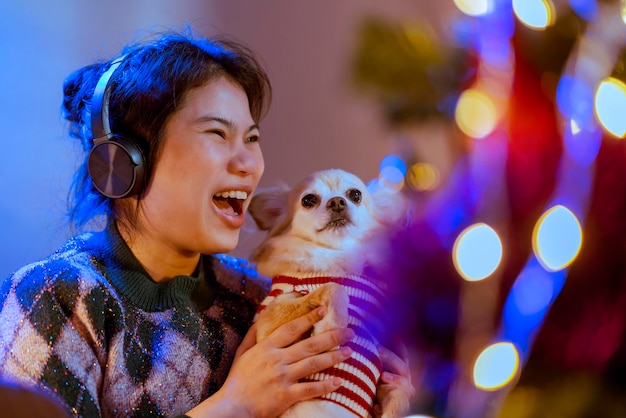 Happiness lifestyle with animal dogfriendly smiling asian\
female woman hold little puppy lap dog while listening music\
headphone wnjoy christmas new year weelend vacation at night\
celebrating herself