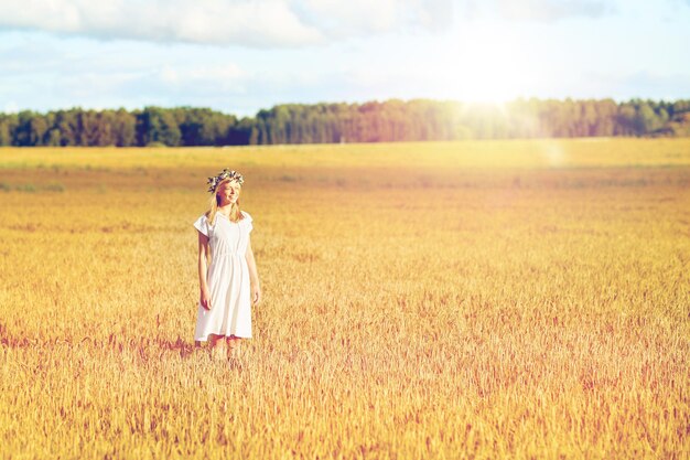 happiness, nature, summer holidays, vacation and people concept - happy smiling young woman or teenage girl in wreath of flowers and white dress on cereal field