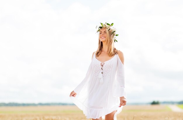 happiness, nature, summer holidays, vacation and people concept - smiling young woman in wreath of flowers and white dress on cereal field