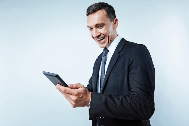 Happiness overload. Waist up shot of an excited mature man wearing a black suit grinning broadly while looking at a screen of a digital tablet over the background.