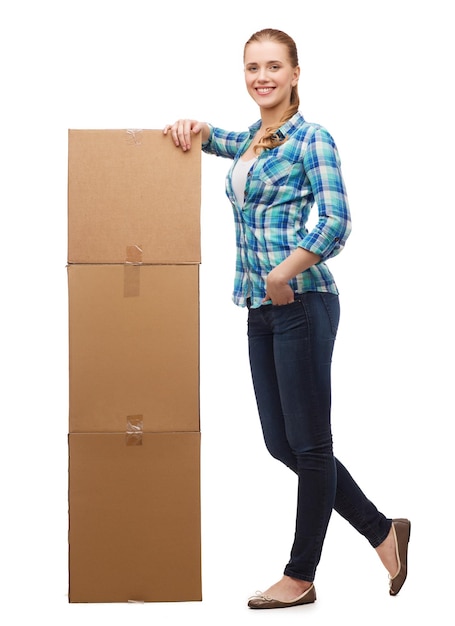 happiness, postal and people concept - smiling young woman standing next to tower of boxes