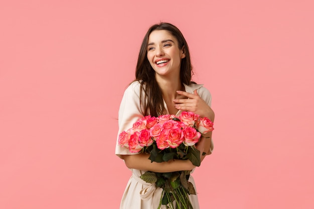 Happiness, romance and beauty concept. Cheerful gorgeous brunette girl in dress, holding roses and smiling flattered, touch chest laughing look away blushing, standing pink wall enthusiastic