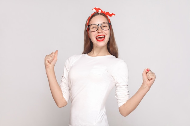 Happiness and victory portrait of beautiful emotional young woman in white t-shirt with freckles, black glasses, red lips and head band. indoor studio shot, isolated on light gray background.