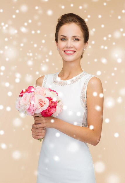 happiness, wedding, holidays, people and celebration concept - smiling woman in white dress with bunch of flowers over beige background and snow