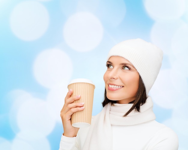 happiness, winter holidays, christmas, beverages and people concept - smiling young woman in white hat and mittens with coffee cup over blue lights background