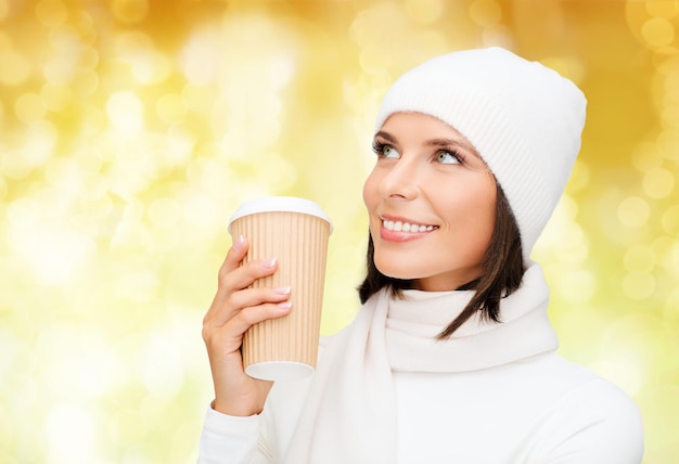 행복, 겨울 방학, 크리스마스, 음료, 그리고 사람들의 개념 - 노란 조명 배경 위에 커피 컵을 든 흰 모자와 장갑을 끼고 웃고 있는 젊은 여성