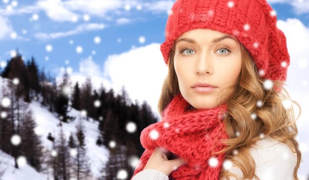 happiness, winter holidays, christmas and people concept - close up of young woman in red hat and scarf over snowy mountains background