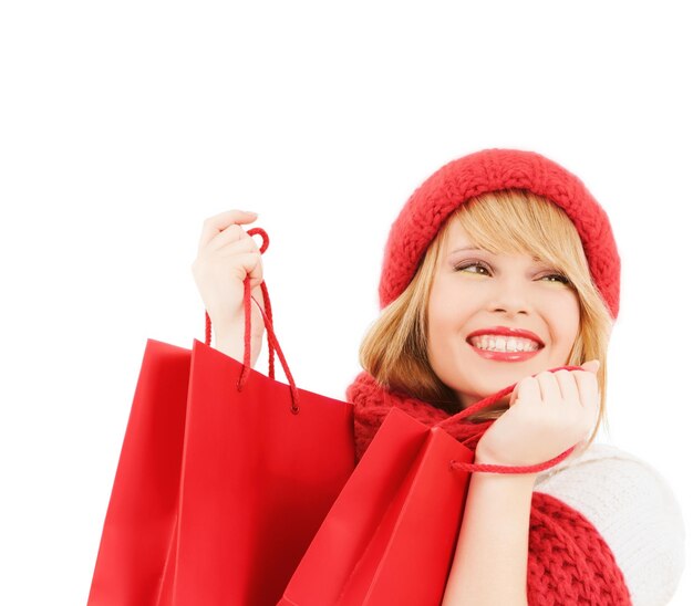 happiness, winter holidays, christmas and people concept - smiling young woman in hat and scarf with red shopping bags over white background