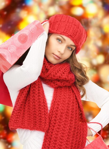 happiness, winter holidays, christmas and people concept - smiling young woman in hat and scarf with shopping bags over red lights background