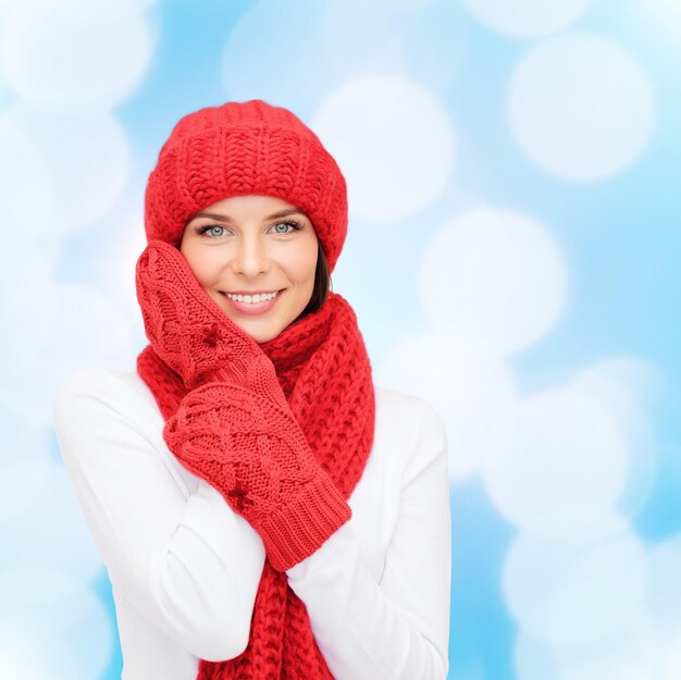 happiness, winter holidays, christmas and people concept - smiling young woman in red hat, scarf and mittens over blue lights background