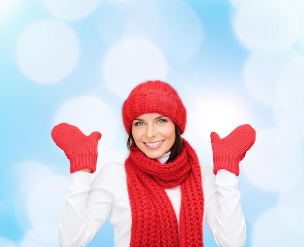 happiness, winter holidays, christmas and people concept - smiling young woman in red hat, scarf and mittens over blue lights background