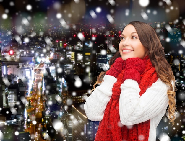 幸福、冬の休日、クリスマスと人々の概念-雪の夜の街の背景に赤いスカーフとミトンの笑顔の若い女性