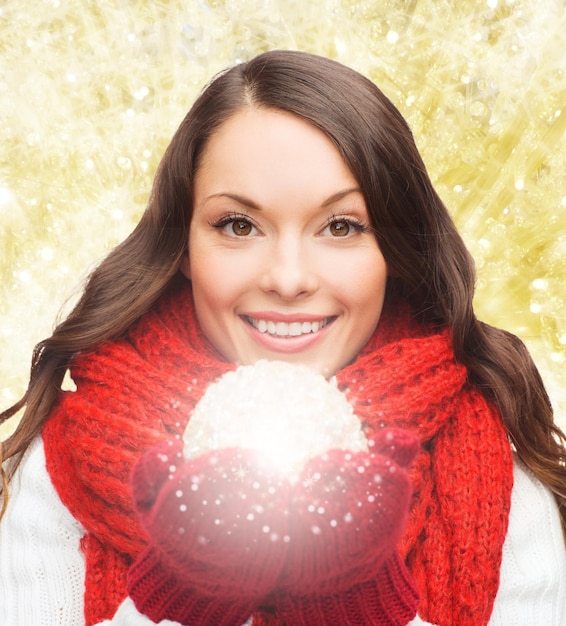 happiness, winter holidays and people concept - smiling young woman in red scarf and mittens with christmas ball over yellow lights background