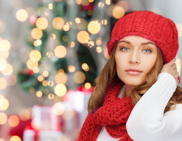 happiness, winter holidays and people concept - young woman in red hat and scarf over christmas tree lights background