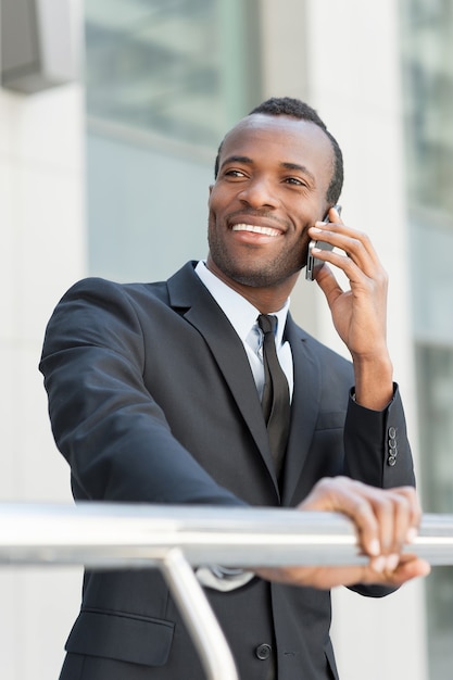 Happy about business going. Handsome young African man in formalwear talking on the mobile phone and smiling while standing outdoors