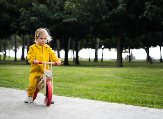 Happy active pretty cute caucasian blonde baby girl kid\
toddlersmiling child about 2 years old wearing bright yellow\
jumpers learning riding run balance bike in summer park\
outdoors