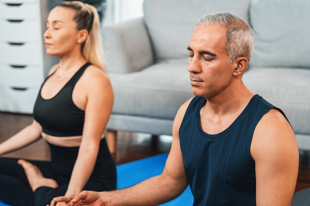 Happy active senior couple in sportswear being doing yoga in meditation posture on exercising mat at home Healthy senior pensioner lifestyle with peaceful mind and serenity Clout