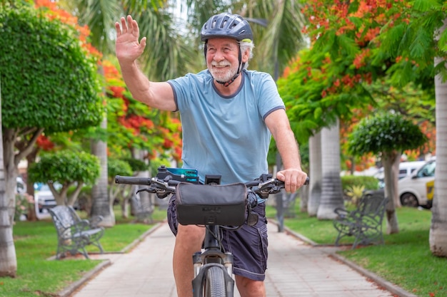 Happy active senior man cycling in the public park with electric bicycle waving hand Concept of healthy lifestyle and sustainable mobility