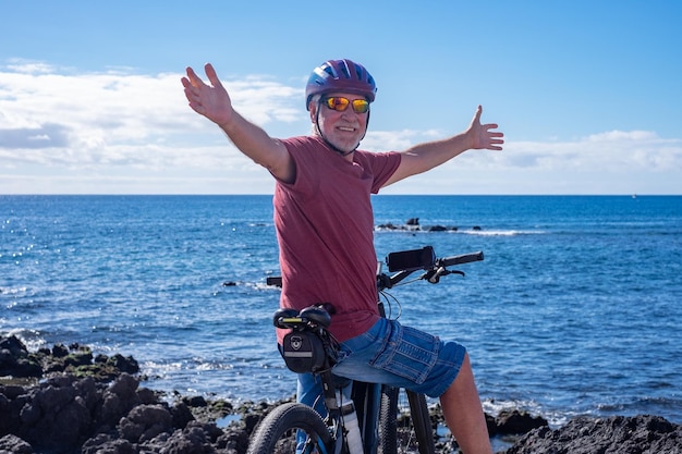 電動自転車でビーチで晴れた日を楽しんでいる両手を広げた幸せなアクティブなシニア男性 本格的な退職後の生活と健康的なライフ スタイルのコンセプト 海の上のホライゾン