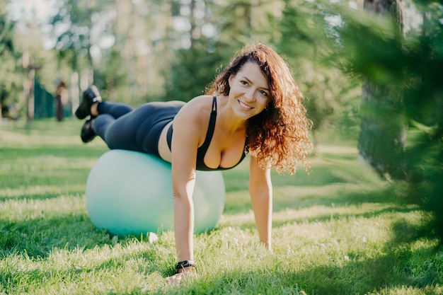 Happy active sportswoman does fitness exercises with gymnastic ball dressed in active wear has workout in open air poses on green grass in forest or park focused somewhere with pleasant smile