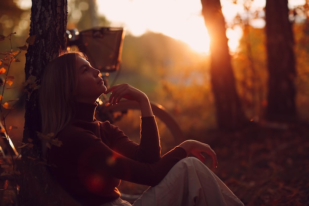 Happy active young woman sitting with vintage bicycle in autumn park at sunset