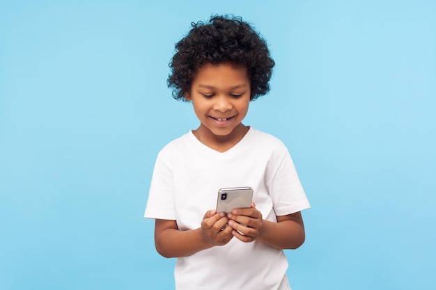 Happy adorable excited little boy with curly hair reading funny message on smartphone and smiling, satisfied with children mobile application, using cellphone. studio shot isolated on blue background