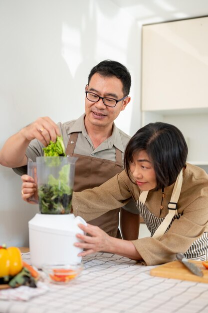 幸せな成人のアジア人カップルがキッチンで一緒にブレンダーを使って健康的な緑色のスムージーを作っています