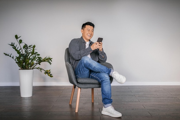 Счастливый взрослый азиатский мужчина печатает на смартфоне, сидя в кресле в минималистском интерьере гостиной, свободное пространство