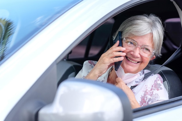 Happy adult attractive caucasian senior woman taking off the\
surgical mask due to coronavirus to answer the mobile phone elderly\
smiling female sitting inside the parked car talking at\
cellphone