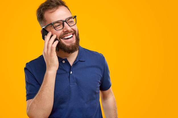 Happy adult bearded male in casual shirt and eyeglasses laughing brightly while receiving good news during phone conversation against yellow background