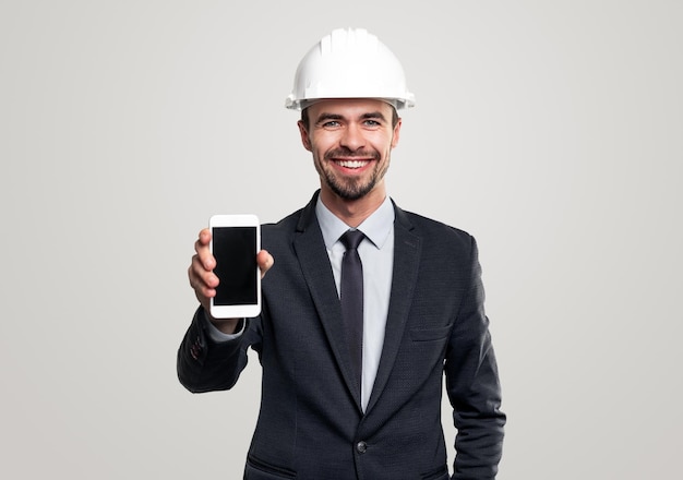 Happy adult bearded male construction engineer in formal suit and hardhat demonstrating mobile phone with blank black screen against gray background