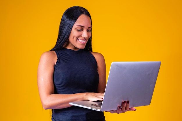Happy adult business lady in elegant dress looking at camera while working on laptop against yellow background