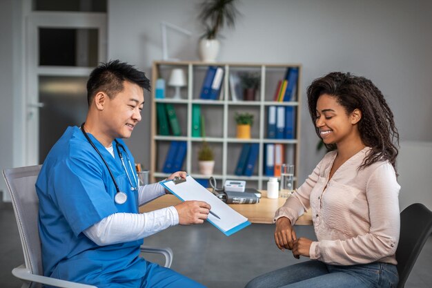 Happy adult chinese man doctor showing contract to young african american female patient in clinic