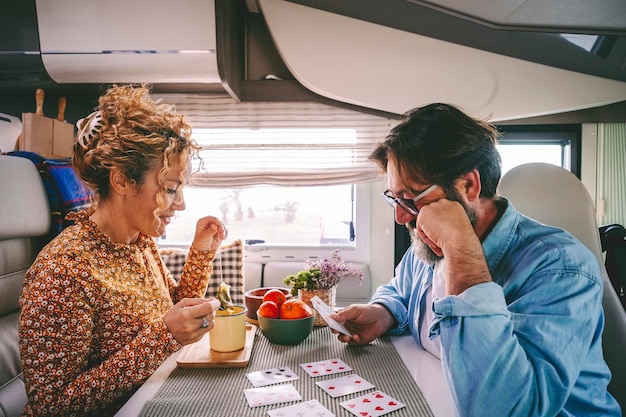 Happy adult couple playing cards during travel camper van\
holiday vacation lifestyle moment enjoying indoor leisure activity\
man and woman sitting at the table van life and renting vehicle for\
trip