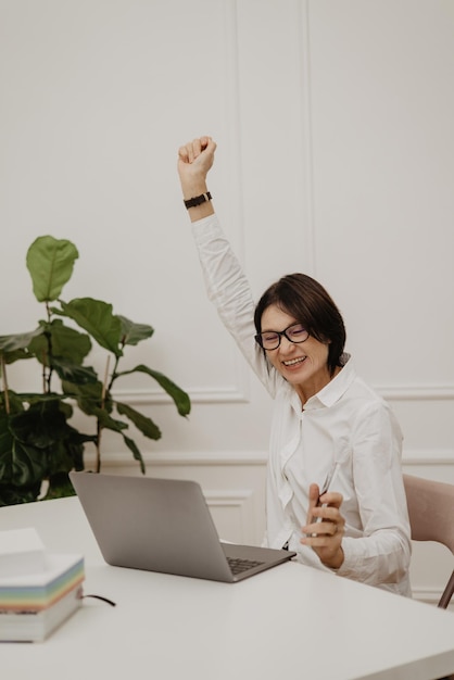 Happy adult european woman closing her eyes rejoices at achievements at work raising hand clenched into fist while sitting at table Success concept