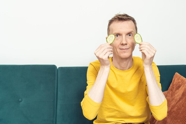 Happy adult man on sofa put patches from fresh cucumber slices on eyes Moisturizing mask for dark circles under eyes