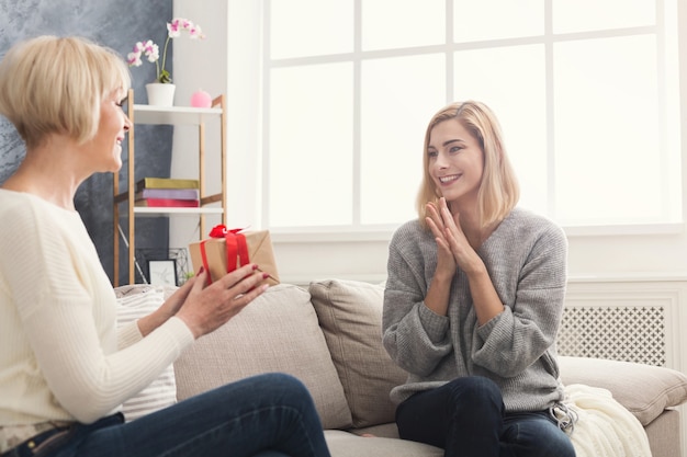 Happy adult mother giving present to surpised daughter . Spending time together, celebrating a holiday at home on weekend. Generation, relationship, celebration concept, closeup