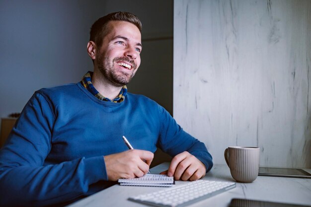 A happy adult student sitting at home office at night and writing down notes from online lecture